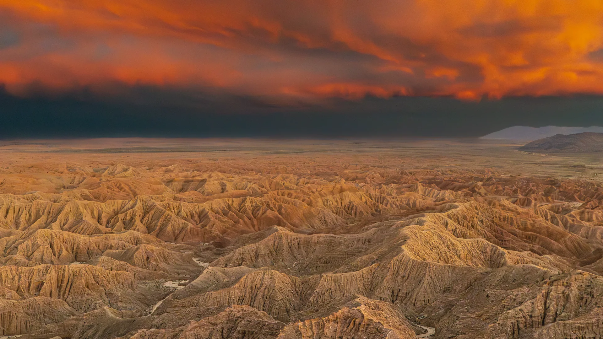 An aerial panorama of deeply eroded badlands terrain glows amber and gold beneath a dramatic storm sky ignited with fiery orange and crimson clouds, the Badlands of the Anza Borrego Desert, where centuries of erosion have carved labyrinthine ridges and ravines stretching toward a distant mountain range on the horizon.