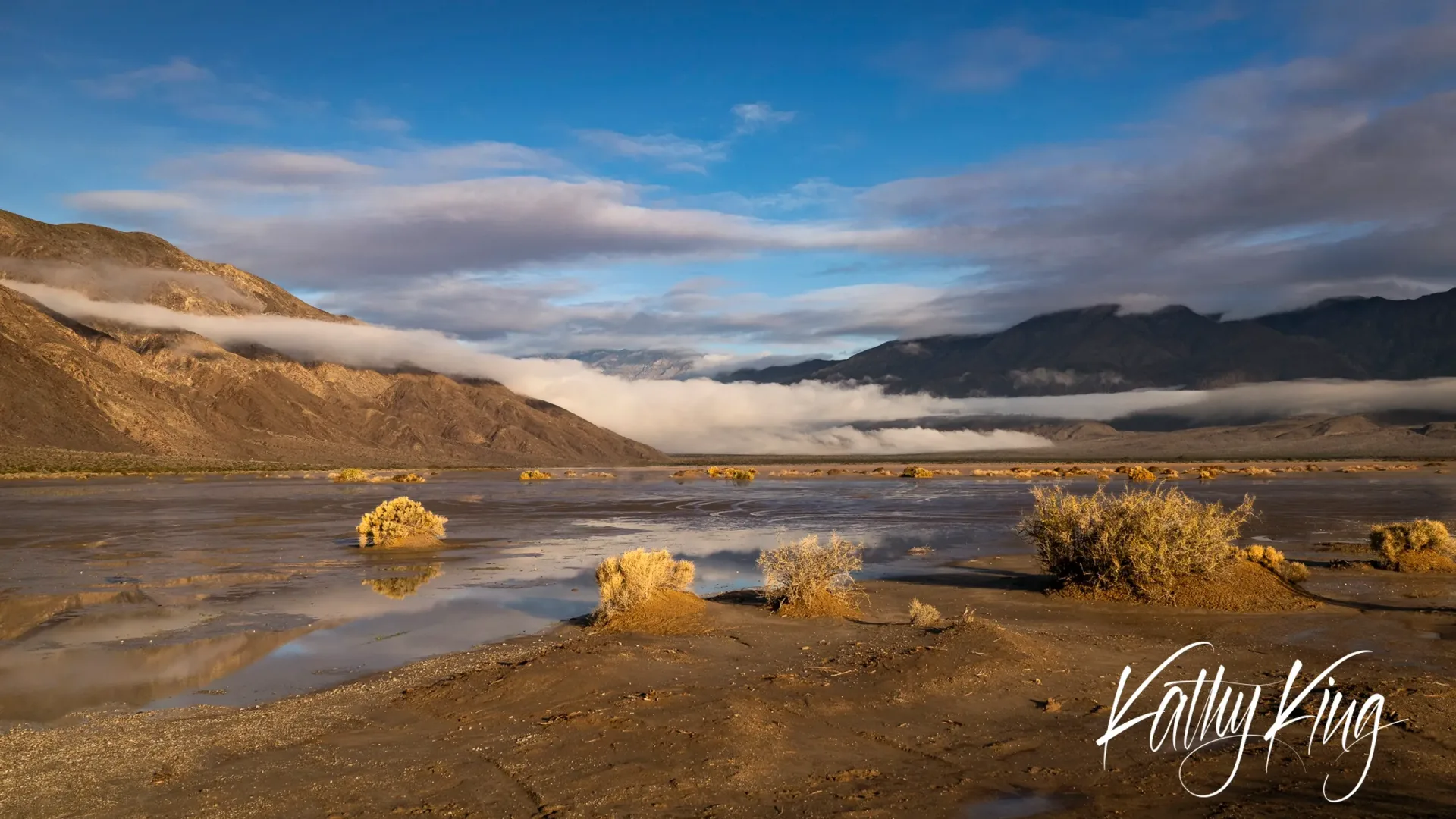 Golden morning light illuminates weathered rock formations scattered across a desert valley floor, while low-hanging clouds drift between distant mountain ranges under a blue sky streaked with wispy clouds. The warm, honeyed light creates a serene and ethereal atmosphere in this expansive wilderness landscape.