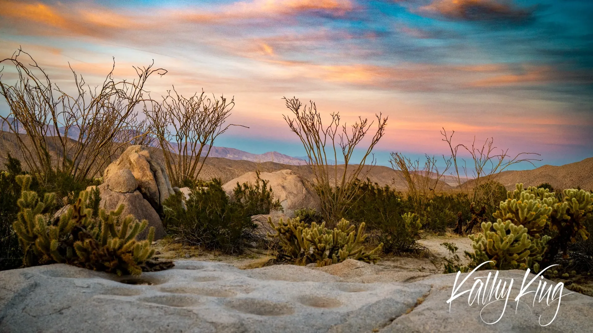 A serene California desert landscape captures the golden hour with dramatic clouds painted in orange and purple hues across the sky. Native desert vegetation including cholla cacti and ocotillo plants frame weathered granite boulders amid rolling hills, creating a peaceful wilderness scene bathed in warm morning light.