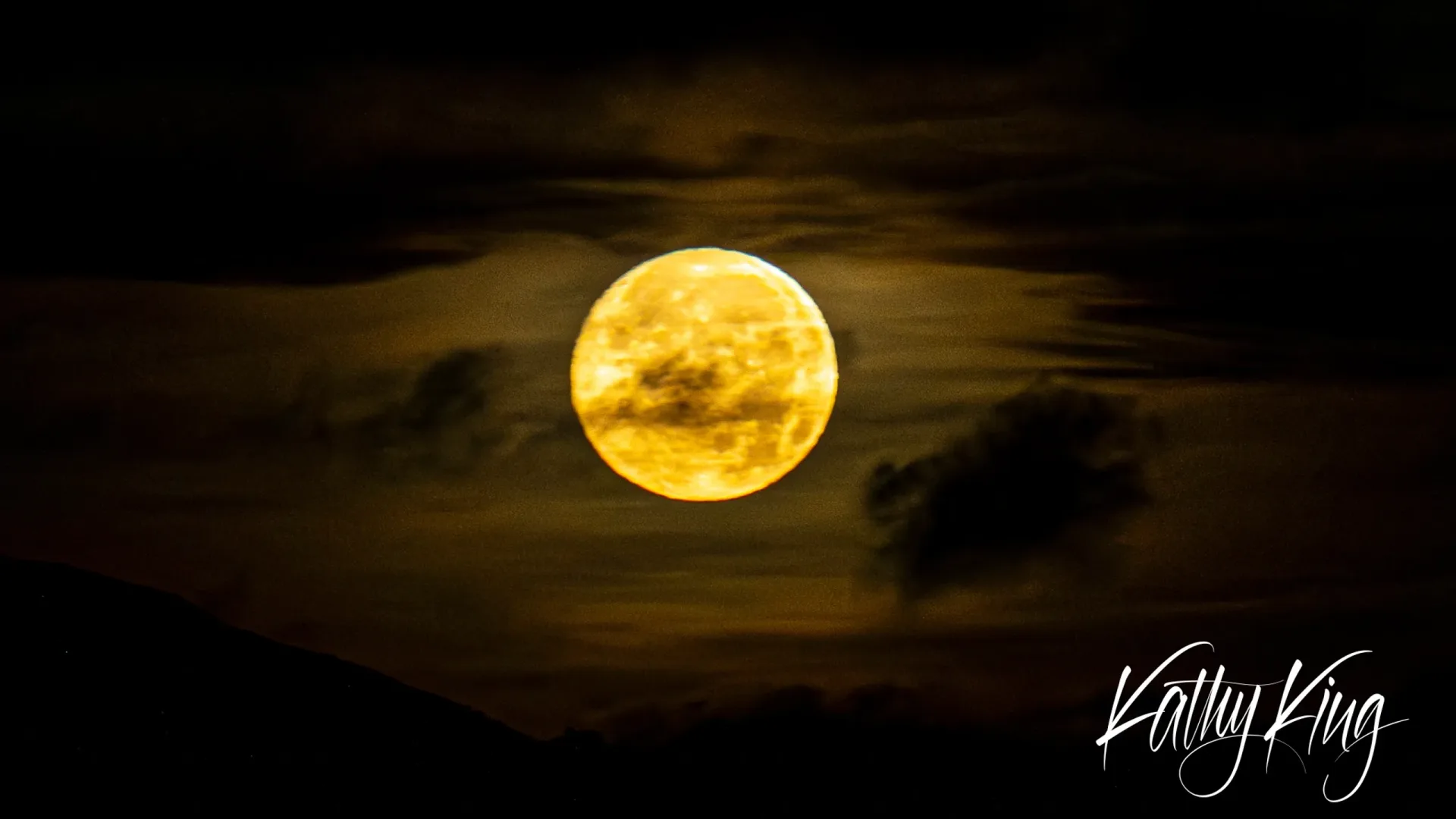 A luminous full moon glows golden yellow against a dramatic night sky filled with dark, drifting clouds. The atmospheric conditions create a warm, amber illumination that contrasts beautifully with the moody cloudscape surrounding the celestial body.
