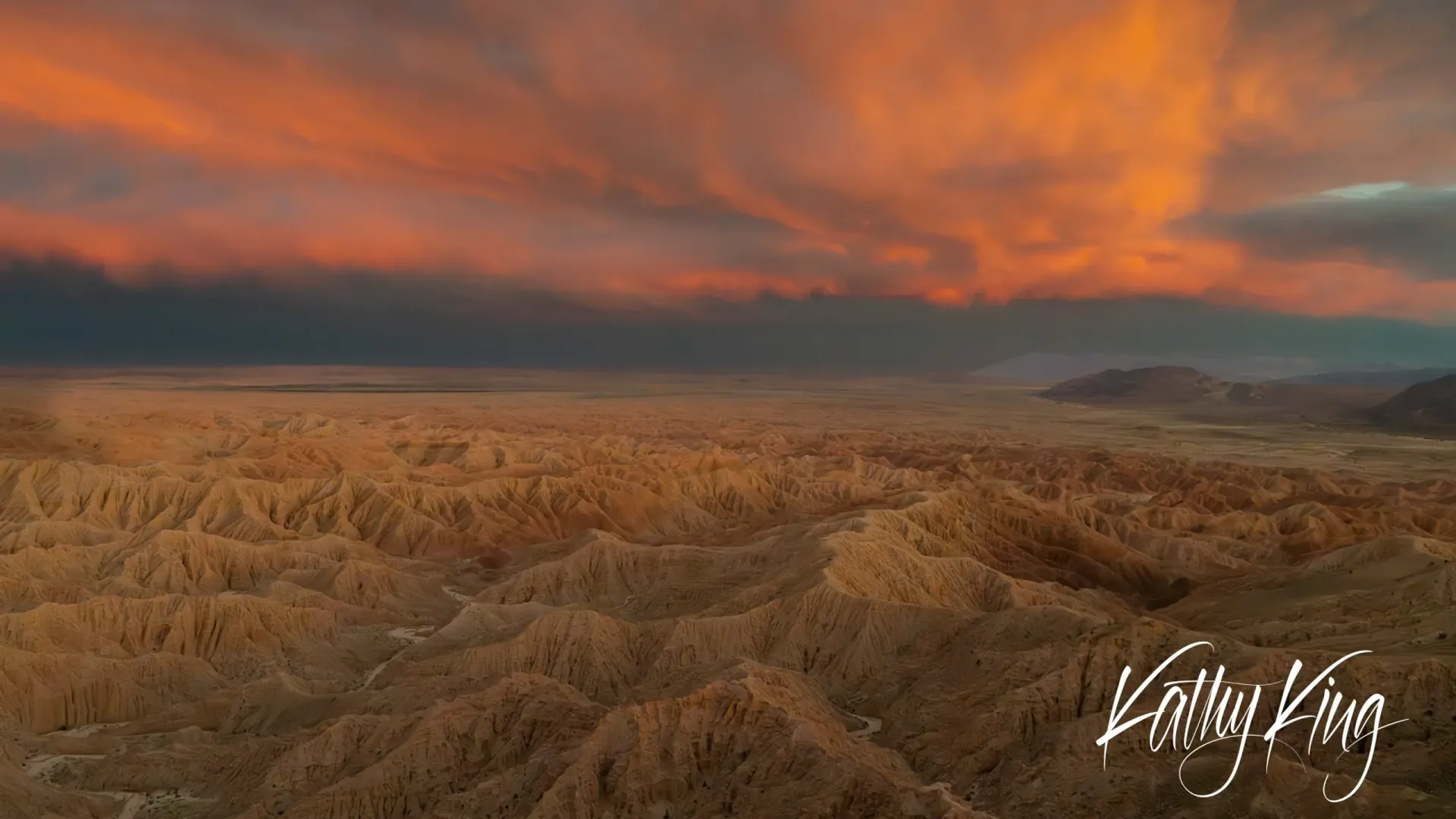 Dramatic orange and red sunset clouds illuminate an expansive desert badlands landscape with deeply eroded ridges and canyons stretching to distant mountains. The warm golden light enhances the rugged terrain's natural sandstone colors, creating a striking contrast against the stormy sky.